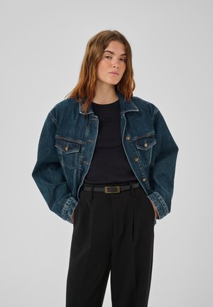 Vrouw met los bruin haar draagt een blauw spijkerjack, zwart shirt en zwarte high-waisted broek met handen in de zakken, neutrale achtergrond.