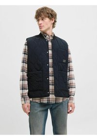 Marineblå quiltet vest med trykknap-åbning, ribbet krave og to frontlommer, båret over en ternet skjorte. Tekstureret materiale og afslappet design.