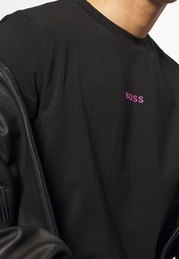 Svart bomullströja med rund hals och ordet "BOSS" i lila broderi, delvis täckt av en svart l äderjacka.