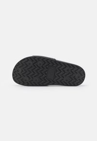 Black rubber sandal sole with textured zigzag pattern and circular grips. Features logo at the center and ridged edges for traction.