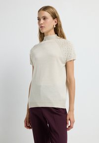 Lichtbeige gebreid top met korte mouwen, hoge halslijn en getextureerde pofdetails op de schouders. Gecombineerd met donkerpaarse broek.