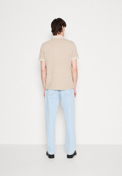 Polo shirt en coton beige à manches courtes, associé à un jean en denim bleu clair. Le tissu semble lisse et le design est simple et ajusté.