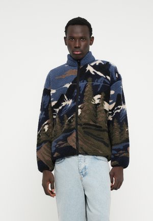 Jachetă din fleece cu un design peisaj montan în nuanțe de albastru, verde și tonuri neutre. Se închide cu fermoar pe toată lungimea, are guler înalt și o textură moale.