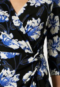 Robe portefeuille noire avec grand motif floral blanc et bleu et manches trois-quarts.