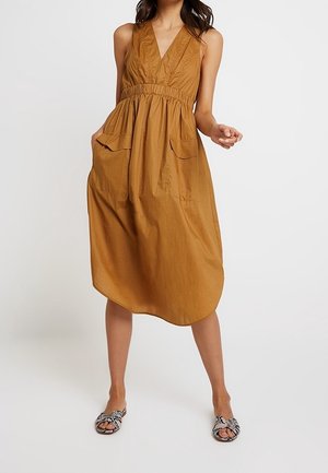 Mouwloze bruine midi-jurk met V-hals en voorzakken, gedragen met zwart-witte sandalen met open tenen en een patroon.