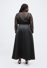 Γυναίκα με πλεξούδες στα μαλλιά, φοράει διαφανές μαύρο πουκάμισο με μακριά μανίκια και μαύρη σατέν μακριά φούστα με ψηλή μέση, φαίνεται από πίσω.