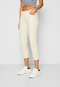 Beige kortere broek met een strakke pasvorm, voorzakken en een klassieke knoopsluiting, gecombineerd met witte sneakers met zwarte accenten.