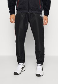 Pantalones deportivos negros con una cinturilla elástica, puños en los tobillos y una textura brillante. Presentan un logo en el muslo y detalles en rojo.