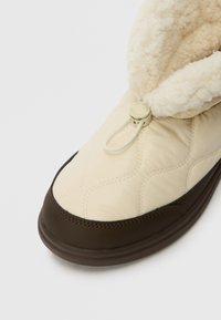 Κρεμ ραμμένος χειμωνιάτικος υπόδημα με σκούρο καφέ σόλα και κάλυμμα δαχτύλου, με επένδυση από φλις και στρογγυλό ρυθμιζόμενο κορδόνι μπροστά.