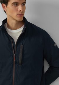 Marineblauwe lichtgewicht jack met hoge kraag, rits aan de voorkant en zijzakken; beschikt over een textuur logo patch en contrasterende rode accenten.