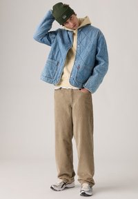 Quilted light blue denim jacket, beige hoodie, beige corduroy pants, gray and black sneakers, worn with a green beanie.