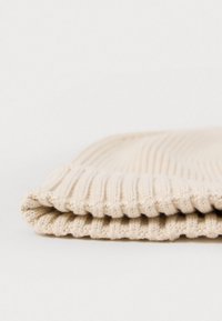 Bonnet en tricot beige clair, avec une texture côtelée et un revers replié. Matériau doux, avec une finition lisse et une légère variation de couleur.