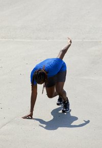 Athlet in einem blauen Sportshirt und schwarzen Shorts bereitet sich auf den Lauf vor, mit einer konzentrierten Haltung und schwarzen Laufschuhen auf einer hellen Fläche.