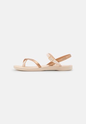 Helle beige Sandalen mit einem goldenen metallischen Kreuzriemen und einem Knöchelriemen, ausgestattet mit einer flachen Gummisohle und einem einfachen Schnallen-Detail.