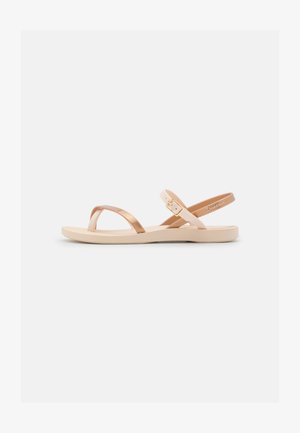 Light beige sandals with a gold metallic cross strap and ankle strap, featuring a flat rubber sole and a simple buckle detail.