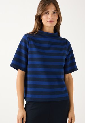 Frau trägt ein blau-schwarzes, horizontal gestreiftes Kurzarmshirt mit hohem Kragen und steht vor einem einfarbigen hellen Hintergrund.