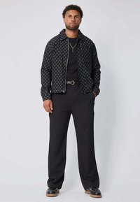 Homme debout avec les mains dans les poches, portant une veste noire à motifs, une chemise noire, un pantalon noir, une ceinture noire et des chaussures noires avec des semelles beiges.