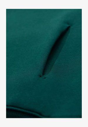 Grüner Stoffpullover mit einer strukturierten Taschenöffnung und glatter Oberfläche, die seine Weichheit und klaren Linien betont.