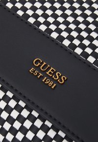 Materiale intrecciato bianco e nero con una base in pelle nera liscia; presenta un logo dorato "GUESS EST 1981".