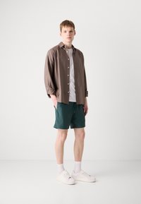 Modèle portant une chemise marron à boutons, un T-shirt blanc, un short vert et des baskets blanches, se tenant devant un fond blanc.