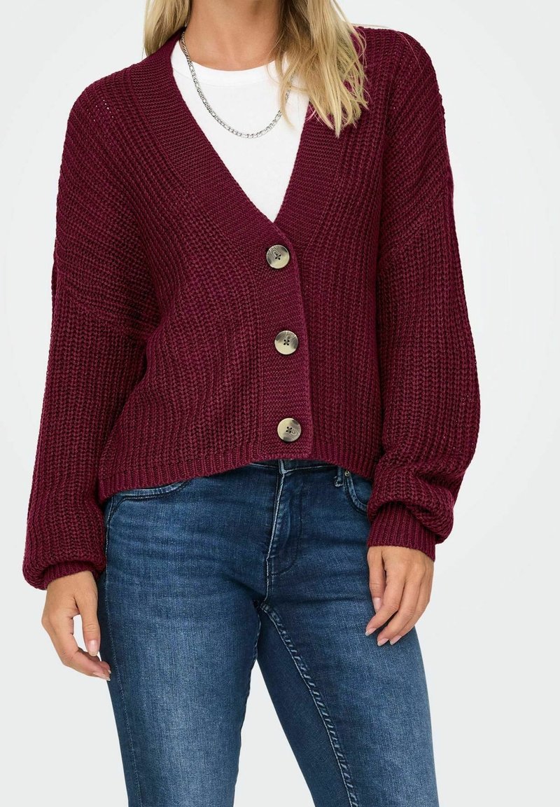 Femme portant un cardigan en maille bordeaux avec de gros boutons sur une chemise blanche et un jean bleu, avec un collier chaîne argenté visible.