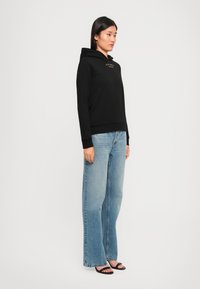 Haut à capuche noir avec une poche avant et un texte en logo doré, associé à un jean évasé bleu clair et des sandales noires à lanières, porté par un mannequin.