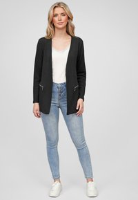 Svart cardigan med en teksturert overflate, åpen front og to glidelås-lommer. Sammen med en hvit cami og lyseblå skinny jeans.