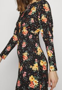 Nainen pukeutuneena mustaan pitkähiharaiseen mekkoon, jossa on oranssia, keltaista ja vaaleanpunaista kukkakuosia, seisoo sivuttain yksinkertaista taustaa vasten.