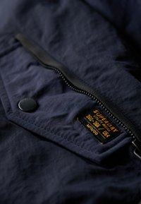 Marineblauwe textuurstof jas met een zichtbare zwarte rits, drukknoop en een label met de merknaam en productdetails.