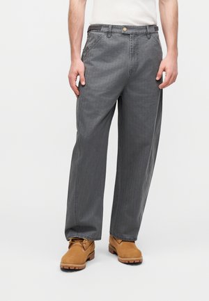 Homme portant un pantalon gris ample avec poches avant et des bottes de travail marron à lacets, debout devant un fond clair uni.