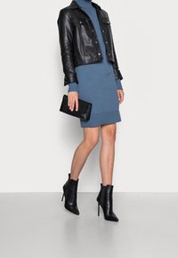 Veste noire en cuir coupée sur une robe en tricot bleue. Pochette noire à la main, associée à des bottines noires à talons pointus. Texture lisse, design ajusté.