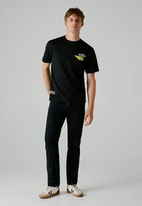 Νεαρός άνδρας που στέκεται φορώντας μαύρο μπλουζάκι με σχέδιο, μαύρα τζιν και λευκά αθλητικά παπούτσια, μπροστά σε ουδέτερο φόντο.