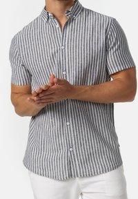 Man in een kortemouw, knoopsluiting shirt met verticale grijs-witte strepen, met de handen voor zijn witte broek gevouwen.