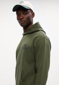 Olívovo zelená mikina s kapucňou a prednou kapsou, s čiernym logom. Skombinovaná s krémovo-zelenou baseballovou čiapkou. Tkanina vyzerá mäkká a texturovaná.