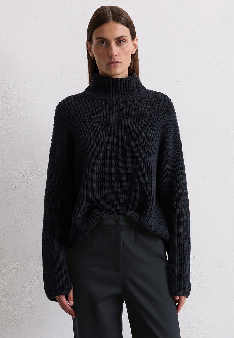 Dark navy high-neck sweater with ribbed texture, oversized fit, and long sleeves, paired with fitted black trousers.