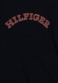 Camiseta de algodón negra con la palabra "HILFIGER" bordada en texto arqueado blanco y rojo en el centro. Textura suave con un acabado jaspeado.