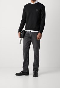 Muž v černé mikině přes bílou košili, šedých džínách a černých mokasínech, držící černou tašku na pozadí s jemnou bílou barvou.