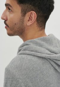 Grå hoodie med en mjuk textur, som har en dragsko vid huven och vinklade sömmar. Öronpiercing syns på subjektets öra.