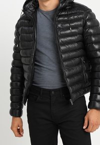 Zwarte leren pufferjack met ritssluiting, horizontale stiksels en een capuchon. Draagt over een grijs T-shirt en zwarte spijkerbroek.