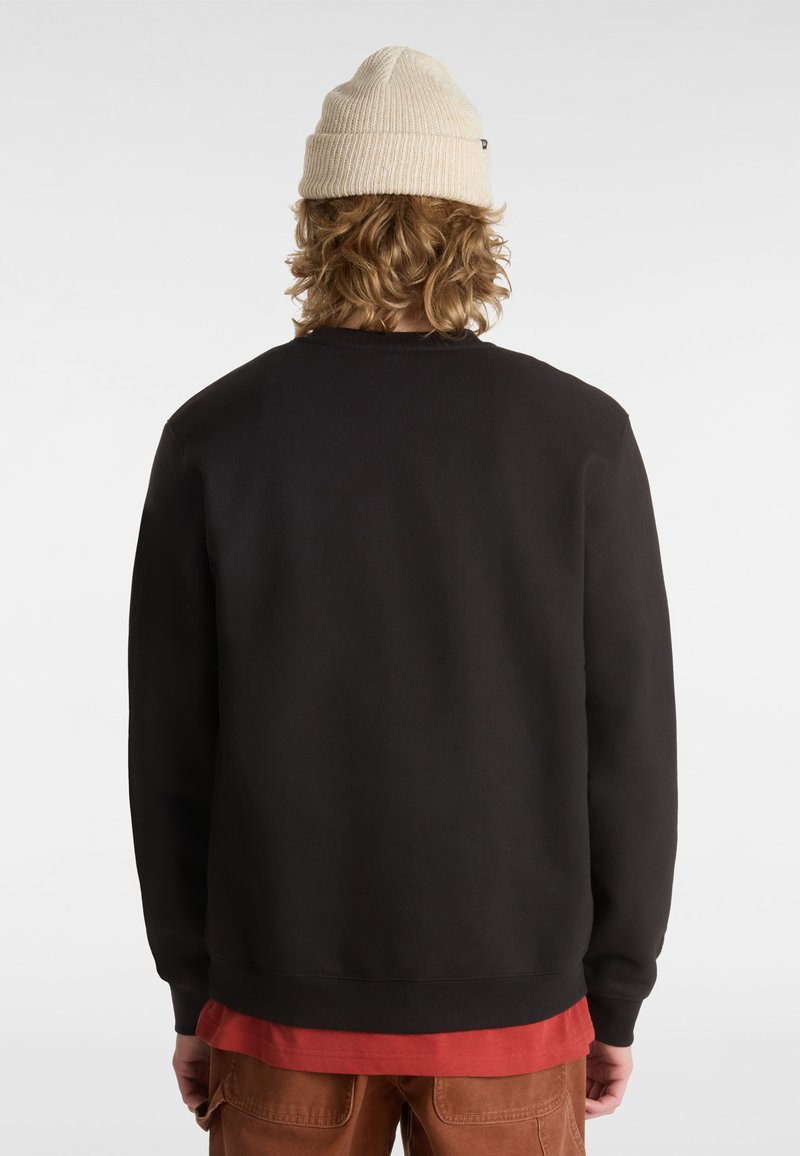 Zwarte sweatshirt met geribde boorden en zoom, gedragen over een rode shirt. De persoon heeft krullend haar en draagt een crèmekleurige beanie.