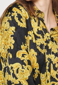 Czarna koszula z guzikami z misternymi żółtymi kwiatowymi i spiralnymi wzorami, noszona przez osobę o jasnej karnacji i brązowych włosach.