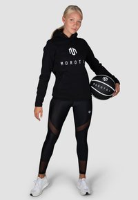 Schwarzer Kapuzenpullover mit weißem "MOROTAI"-Logo, kombiniert mit schwarzen Leggings mit Netzpaneelen und weißen Sportschuhen. Hält einen schwarzen Basketball.