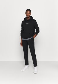 Black pullover hoodie with a kangaroo pocket and a graphic text design, paired with black tapered pants and white sneakers, standing on a plain background.