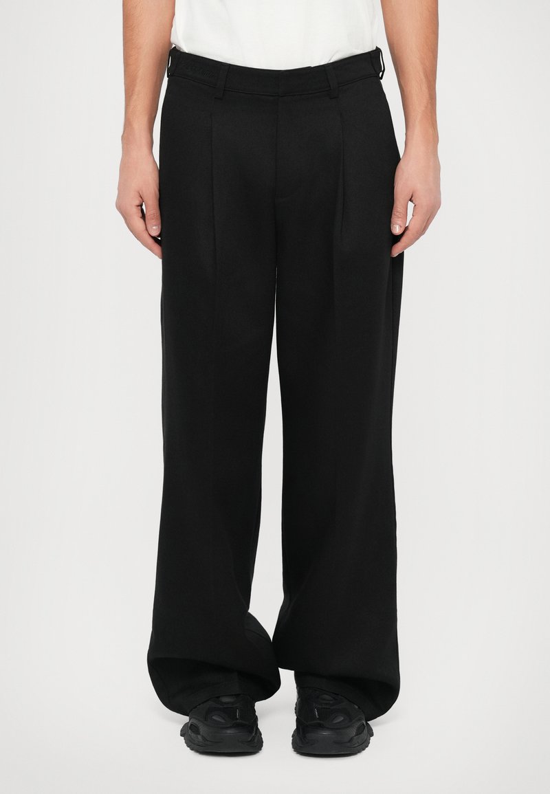 Schwarze weit geschnittene Hose aus glattem Stoff, mit vorderen Falten, Gürtelschlaufen und taillierter Passform. Kombiniert mit schwarzen Sneakern.