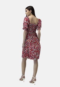 Vestido rojo de flores con mangas abullonadas, una cintura fruncida y un escote cuadrado. La tela presenta patrones vibrantes de múltiples colores. Combinado con tacones blancos.