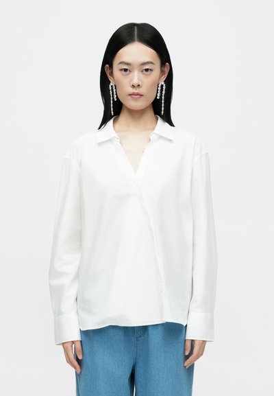 Chemise blanche à col, à manches longues et à coupe décontractée. Tissu en coton lisse. Portée avec un pantalon en denim bleu.
