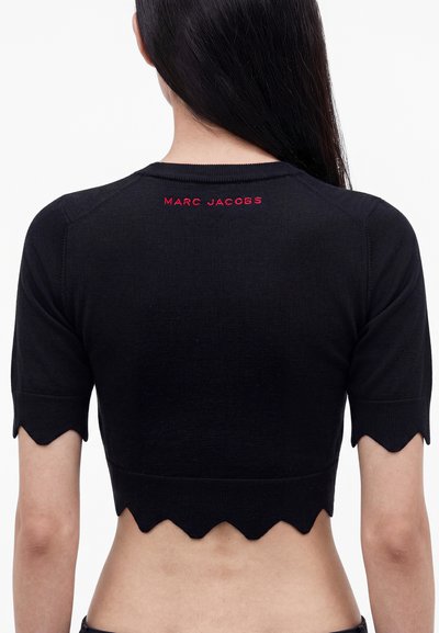Άτομο με μακριά σκούρα μαλλιά φοράει μαύρο crop top με κοντό μανίκι, κυματιστές άκρες και το "MARC JACOBS" με κόκκινα γράμματα στο πάνω μέρος της πλάτης.