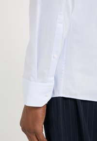 Gros plan sur le bras d'une personne portant une chemise à manches longues texturée bleu clair avec un poignet à boutons et un pantalon sombre à fines rayures.