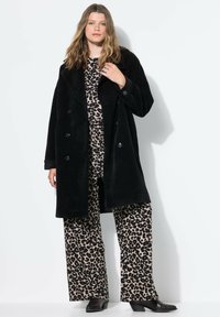 Černý, oversized kabát z texturované látky s velkými knoflíky, vrstvený přes béžový a černý leopardí potisk a černé kotníčkové boty.