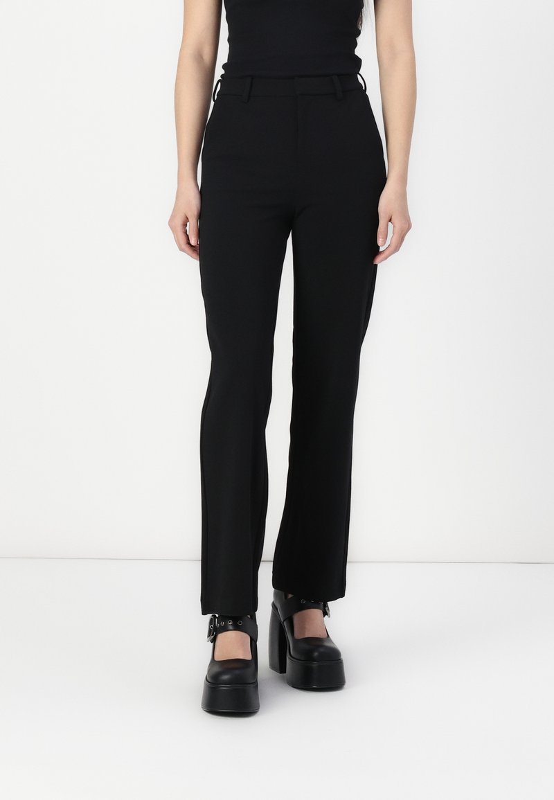 Femme portant un pantalon noir ajusté et des chaussures Mary Jane à plateforme noires avec des brides à boucle, debout sur un fond blanc.